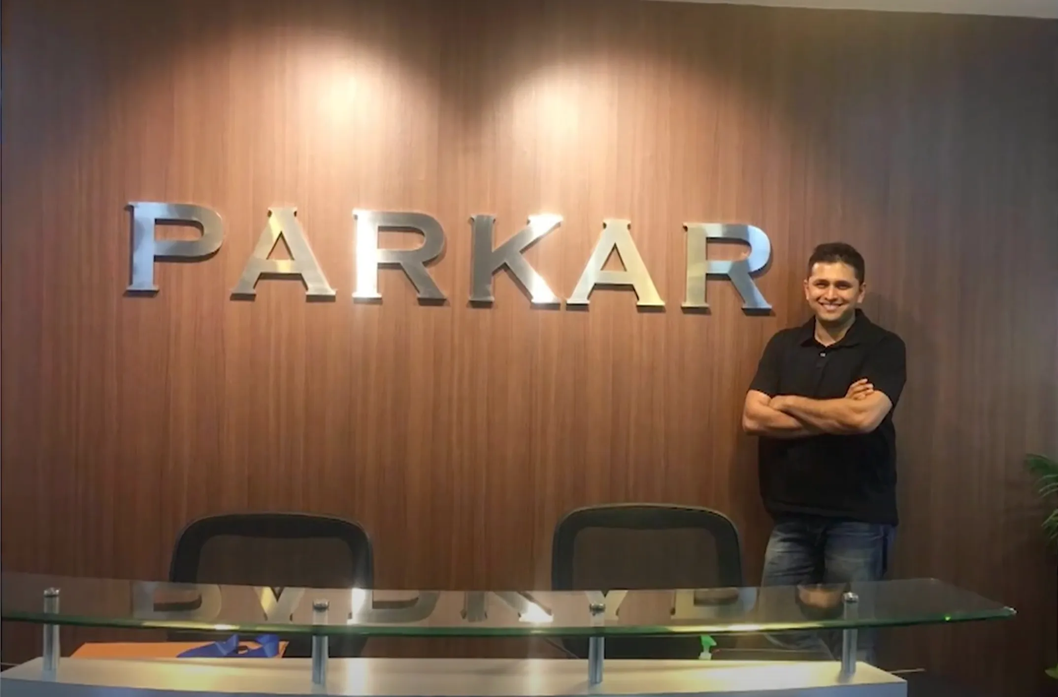 Man in a black shirt standing with arms crossed beside a reception desk under a metallic PARKAR sign.