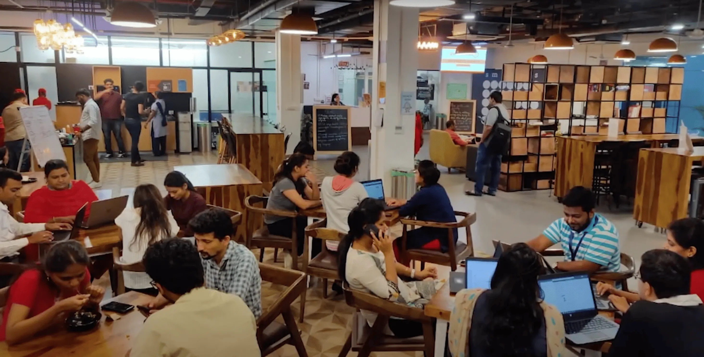 Busy coworking space with people working on laptops at wooden tables.