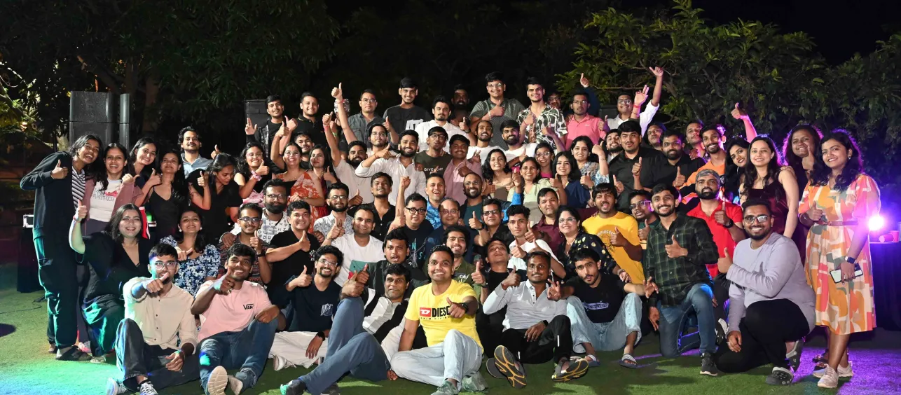 Large group of diverse young adults gathered outdoors at night, smiling and giving thumbs up.