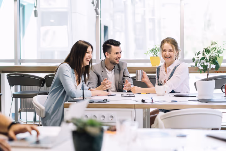 Three young colleagues sitting at a table in a bright office, smiling and engaged in conversation.